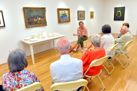 A group of people are sat in a row in front of paintings within the gallery and a table of white ceramic crockery. The artist is sat in front of the group, she has her hands out and is laughing openly