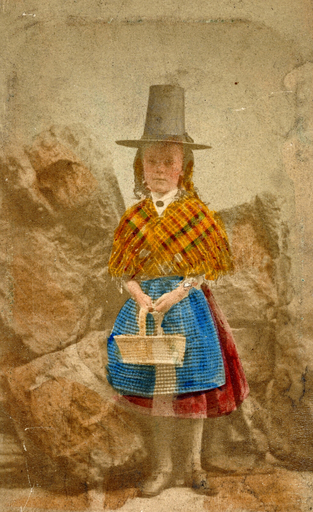 An old colourised photograph of a girl in a tall Welsh hat and a shawl, holding a basket.
