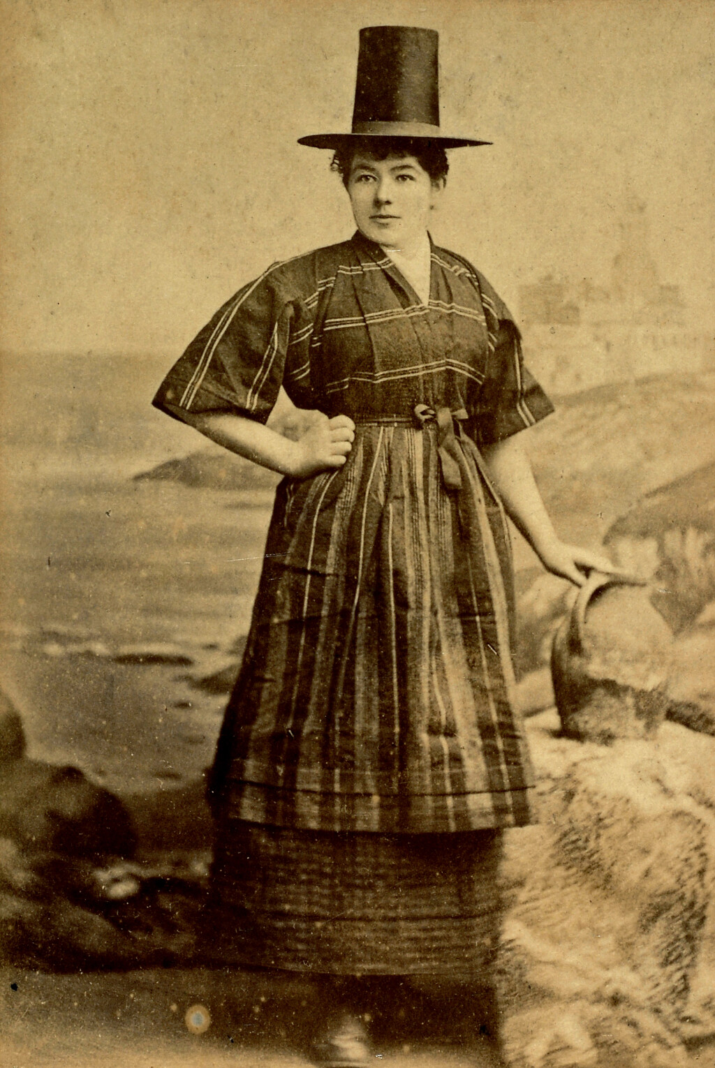 A sepia photograph of a woman wearing a tall Welsh hat, standing against a background of a coastal scene, with one hand on her hip and the other resting on a basket.